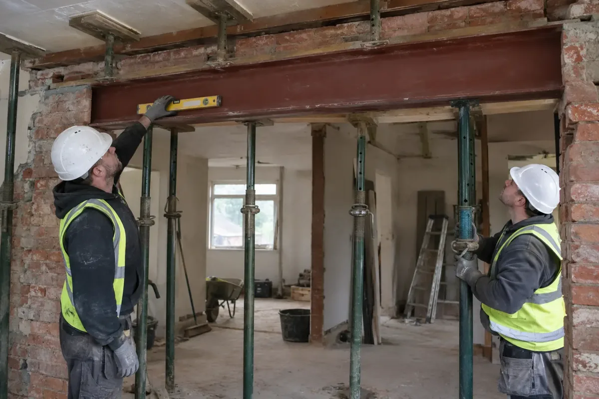 Two builders with helmets and gloves checking a newly installed steel lintel across a large internal opening in a formerly load-bearing brick wall. Clearly visible correctly arranged temporary steel props support remaining structure, engineers' level, no unsupported masonry.